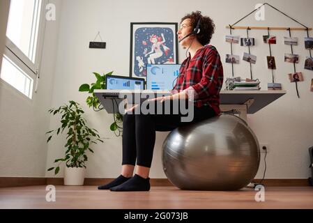 Frau mit Telearbeit, die vor ihr auf einem Fitball sitzt Schreibtisch, der über ein Headset spricht Stockfoto