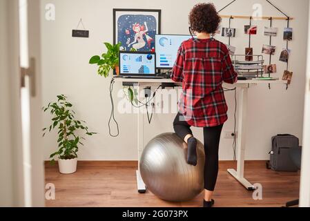 Frau, die von zu Hause aus mit einem höhenverstellbaren Schreibtisch steht und ihr Knie auf einem Fitball ruht Stockfoto