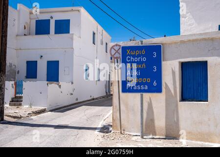 Griechenland, Kimolos-Insel. 18.Mai 2021. Straßenschild zweisprachig, griechisch und englisch auf Stange. Konzept des Reiseziels. Traditionelle griechische kykladische Gebäude Stockfoto