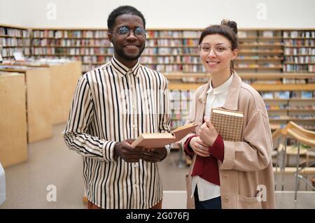 Portrait von zwei Studenten, die Bücher halten und die Kamera anlächeln, während sie in der Bibliothek stehen Stockfoto