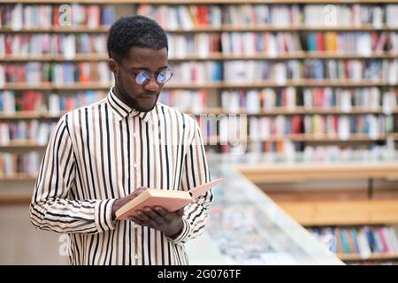 Afrikanischer junger Mann in einer Brille, der das Buch in der Bibliothek untersucht Stockfoto