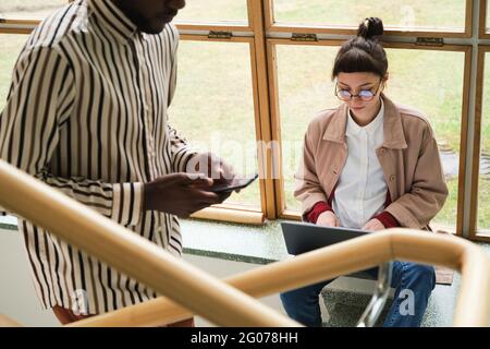 Afrikanischer Mann, der im Hintergrund im Korridor ein Mobiltelefon benutzt und die Treppe hinauf bewegt, während eine Frau am Laptop arbeitet Stockfoto