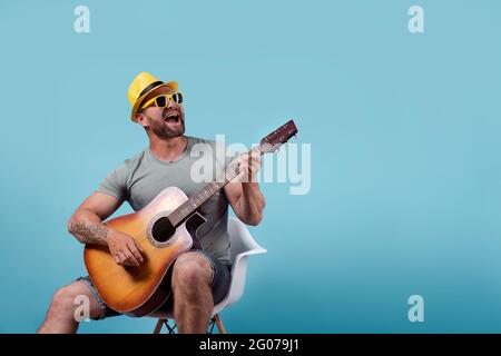 Schöner bärtiger Gitarrist in gelbem Hut und Sonnenbrille, der auf einem Stuhl sitzt und singt, während er auf akustischer Gitarre spielt, isoliert auf blauem Hintergrund Stockfoto