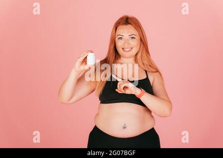 Fröhliche Plus-Größe Frau trägt sportliche Outfit zeigt auf Glas Pillen, während auf rosa Hintergrund stehen. Stockfoto