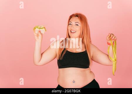 Fröhliche Frau in Übergröße, die ein sportliches Outfit mit Fast Food Burger und Maßband trägt, während sie auf pinkem Studiohintergrund steht. Stockfoto