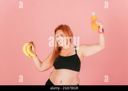 Glückliche übergroße Frau trägt sportliche Top hält Haufen Bananen und eine Flasche Detox Saft, während sie auf rosa Studio Hintergrund stehen. Stockfoto