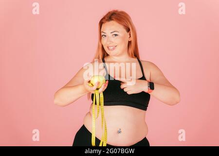 Porträt einer schönen übergroßen Frau im Sportoutfit, die Apfel mit Maßband hält, mit dem Finger zeigt und lächelt, während sie auf die Kamera schaut. Stockfoto