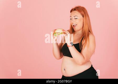 Glücklich Rotschopf Frau mit plus size Körper essen Fast Food Burger, während auf rosa Hintergrund stehen. Stockfoto