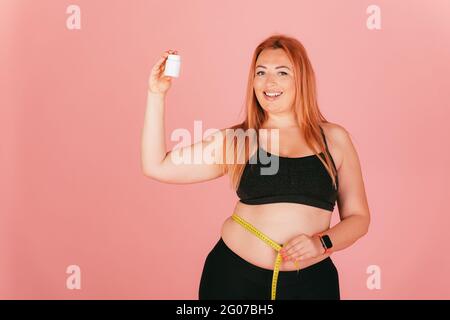 Fröhliche Plus Größe Frau trägt sportliche Outfit Messung ihrer Taille und hält Glas von Pillen, während auf rosa Hintergrund. Stockfoto