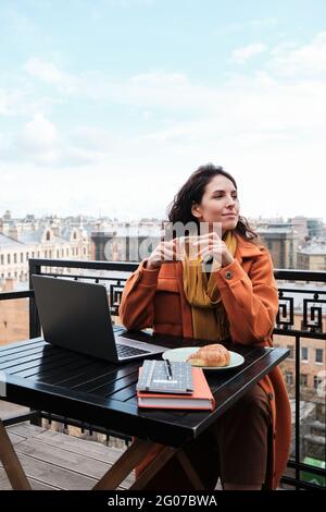 Junge Frau, die mit einem Laptop am Tisch sitzt und die Tasse Tee genießt und die Aussicht betrachtet Stockfoto