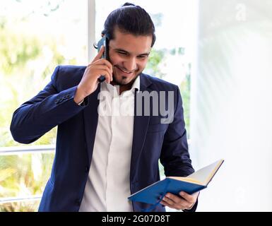 Lächelnder junger Latino-Geschäftsmann in einem Telefonanruf und Lesen auf dem Notizblock. Home Office-Konzept Stockfoto