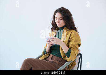 Junge Frau, die auf einem Stuhl sitzt und vor dem weißen Hintergrund ein Mobiltelefon benutzt Stockfoto