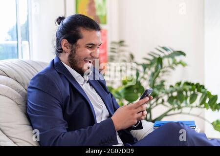 Junger Latino-Mann mit langen Haaren und Piercings in Anzug lächelnd gekleidet, während SMS auf dem Smartphone im Home Office. Home Office-Konzept Stockfoto