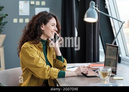 Junge lächelnde Frau, die am Tisch sitzt und am Laptop arbeitet und zu Hause auf dem Mobiltelefon spricht Stockfoto