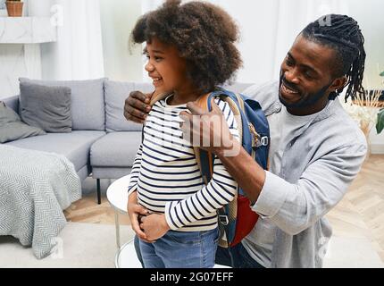 Glücklicher afroamerikanischer Vater hilft der Tochter, sich für die Schule vorzubereiten und legt ihr eine Schultasche auf den Rücken Stockfoto