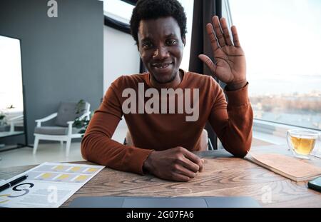 Porträt eines afrikanischen Geschäftsmannes, der die Kamera anlächelt und seine Hand schwenkt, während er im Büro sitzt Stockfoto