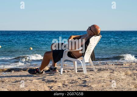 Eilat, Israel - 24. Mai 2021: Ein Mann, der sich an einem klaren, sonnigen Tag in einem Plastikstuhl an einem Strand in Eilat, Israel, entspannt Stockfoto