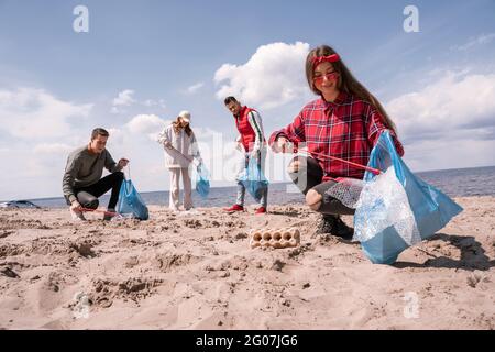 Lächelnde junge Frau, die Müllbeutel in der Hand hält und Müll mit einem Grabber auf Sand in der Nähe einer Gruppe von Freiwilligen sammelt Stockfoto