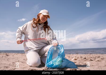 Glückliche Frau in der Mütze, die Müllbeutel hält und Müll auf Sand sammelt Stockfoto