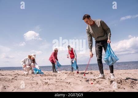 Glücklicher Mann, der Müllbeutel in der Hand hält und Müll auf Sand in der Nähe einer Gruppe von Freiwilligen aufsammelt Stockfoto