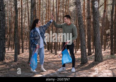 Die ganze Länge des glücklichen Paares mit Mülltüten geben hohe fünf beim Aufnehmen von Müll mit Greifwerkzeugen im Wald Stockfoto