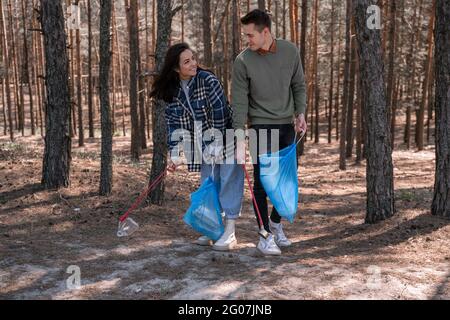 Glückliche junge Frau und Mann mit Mülltüten, die mit Greifwerkzeugen im Wald Müll aufsammeln Stockfoto