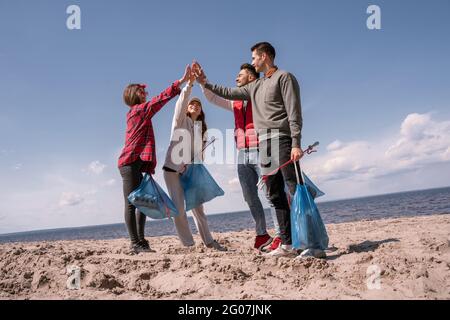 Eine Gruppe von fröhlichen Freiwilligen, die Müllsäcke in der Hand halten und fünf hoch geben Stockfoto