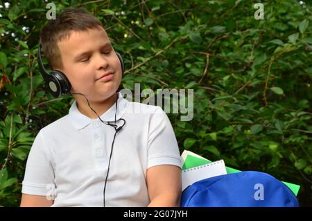 Ein Schuljunge in einem weißen Hemd neben einer Aktentasche im Park spielt Musik auf Kopfhörern. Schüler lernen Hörübungen in einem Park Stockfoto