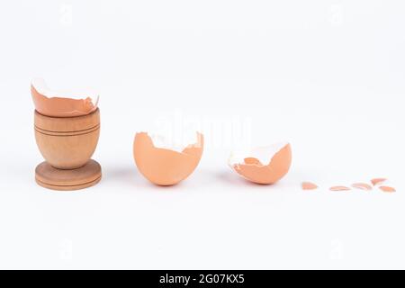 Horizontale Aufnahme einer zerbrochenen Eierschale in einem alten hölzernen Eierständer und mehreren anderen zerbrochenen Schalen auf einer weißen Oberfläche. Minimalismus. Konzeptkunst für Essen o Stockfoto