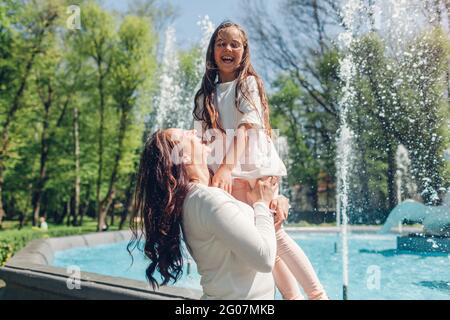 Junge Mutter und ihre Tochter haben Spaß am Brunnen im Sommerpark. Frau hält Mädchen. Familie lacht, Spiele spielen Stockfoto