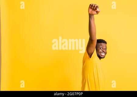 Glücklicher junger afroamerikanischer Mann in gelbem trendigen T-Shirt, mit Siegesgeste, die Hand nach oben bewegen. Fröhlich lächelnder schwarzer Tausendjähriger Mann Stockfoto