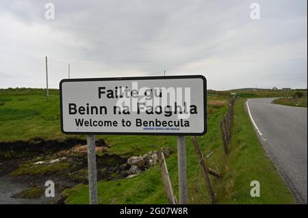 Willkommen bei Benbecula Zeichen in Englisch und Schottisch Gälisch. Verschwommener ländlicher Hintergrund mit Straße. Bewölktes Wetter. Äußere Hebriden, Schottland. Stockfoto