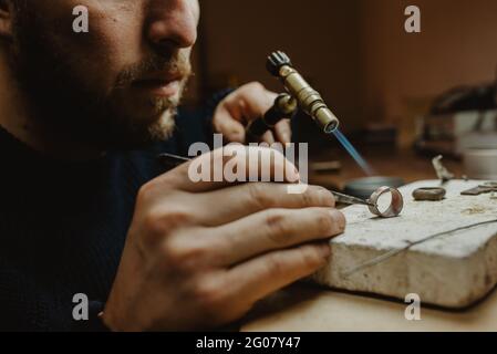 Goldschmied verwendet eine Lötlampe, um beim Herstellen kleine Metallornamente zu erhitzen Schmuck auf der Werkbank Stockfoto