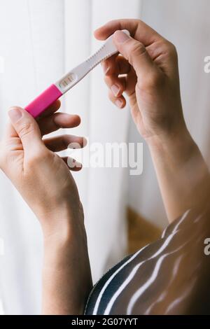 Beschnittene, nicht erkennbare Hände einer Frau, die neben einem Fenster einen Schwangerschaftstest durchgeführt hat Stockfoto