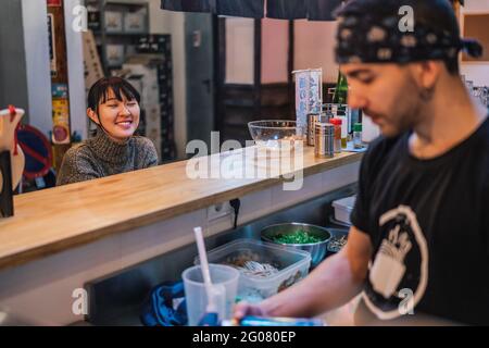 Asiatische Frau in legerer Kleidung sitzt an der Theke und spricht mit männlichen Arbeiter der modernen Ramen-Bar Stockfoto
