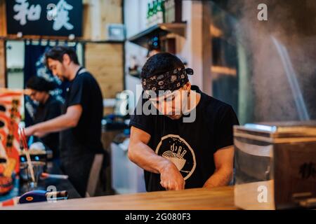 Junger Mann auf der Schürze kocht asiatische Gerichte, während er steht Theke in Ramen Bar Stockfoto
