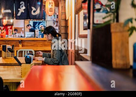 Junge asiatische Frau im Pullover, die an der Holztheke im Café isst Stockfoto
