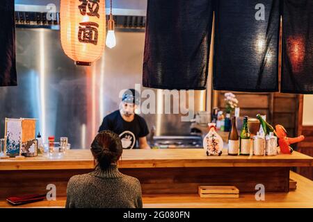 Rückansicht einer gesichtslosen, schwarzhaarigen Frau im Pullover, die an der Theke in der gemütlichen Ramen-Bar sitzt Stockfoto