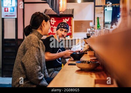 Seitenansicht von Frau und Mann, die miteinander kommunizierten, während sie an der Holztheke im modernen Café asiatisches Essen aßen Stockfoto