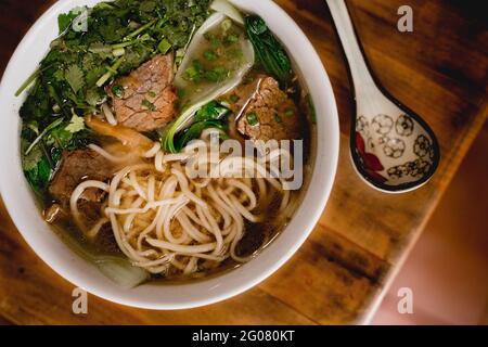 Von oben große weiße Keramikschüssel mit Rinderramen-Suppe Und bunten Löffel auf Holztisch platziert Stockfoto