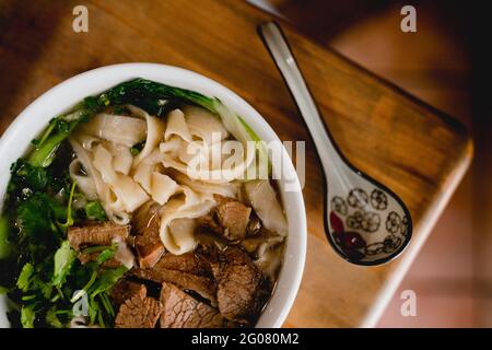 Von oben große weiße Keramikschüssel mit Rinderramen-Suppe Und bunten Löffel auf Holztisch platziert Stockfoto