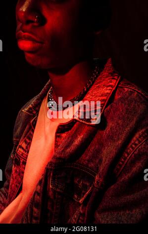 Crop Calm stilvoller afroamerikanischer Mann in Jeans-Jacke unter Neonrotes Licht im Schatten auf schwarzem Hintergrund Stockfoto
