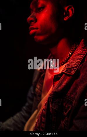 Crop Calm stilvoller afroamerikanischer Mann in Jeans-Jacke unter Neonrotes Licht im Schatten auf schwarzem Hintergrund, das wegschaut Stockfoto