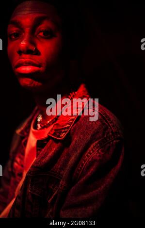 Crop Calm stilvoller afroamerikanischer Mann in Jeans-Jacke unter Neonrotes Licht im Schatten auf schwarzem Hintergrund Kamera Stockfoto