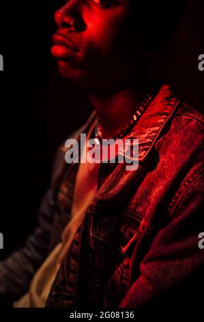 Crop Calm stilvoller afroamerikanischer Mann in Jeans-Jacke unter Neonrotes Licht im Schatten auf schwarzem Hintergrund, das wegschaut Stockfoto