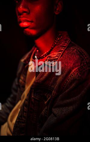 Crop Calm stilvoller afroamerikanischer Mann in Jeans-Jacke unter Neonrotes Licht im Schatten auf schwarzem Hintergrund Kamera Stockfoto