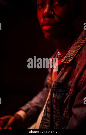 Crop Calm stilvoller afroamerikanischer Mann in Jeans-Jacke unter Neonrotes Licht im Schatten auf schwarzem Hintergrund Kamera Stockfoto