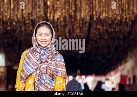 Junge östliche Frau in traditioneller Kleidung und Kopftuch lächelnd und schauend auf die Kamera, während sie vor dem verschwommenen Hintergrund des alten manama souq baz steht Stockfoto