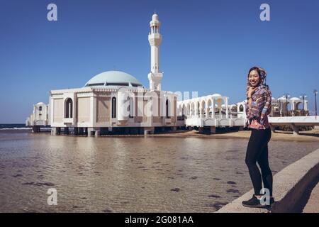 Seitenansicht einer jungen Frau im traditionellen Kopftuch, die in der Nähe des Wassers gegen die berühmte schwimmende Moschee oder die Al Rahma Moschee mit weißen Steinmauern in Jedd steht Stockfoto