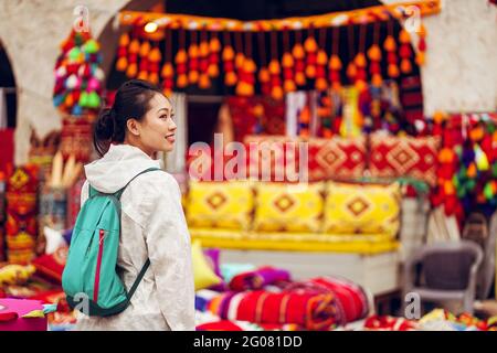 Rückansicht Zufriedene asiatische weibliche Touristin mit Rucksack lächelnd weg Während Sie sich gegen verschiedene traditionelle orientalische Kleidung und Souvenirs wehren Stockfoto